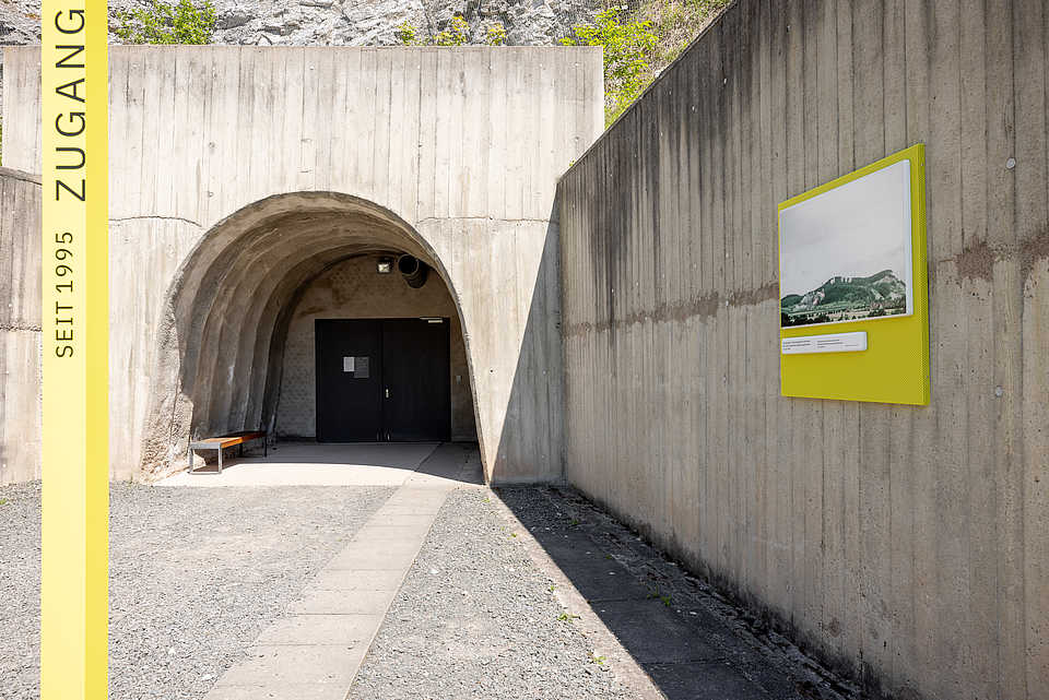 Eingangsbereich zur Stollenanlage der KZ-Gedenkstätte Mittelbau-Dora mit Betonmauer, Infotafel und dem gelben Schriftzug ‚Zugang seit 1995‘.