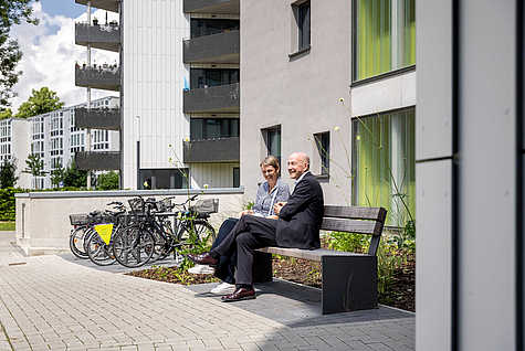 Zwei Personen, eine Frau und ein Mann, sitzen auf einer Bank vor einem modernen Wohngebäude. Die Frau trägt eine graue Jacke und weiße Schuhe, während der Mann einen dunklen Anzug trägt. Hinter ihnen sind mehrere Fahrräder in einem Fahrradständer abgestellt. Das Gebäude hat graue Wände und grüne Akzente, mit Balkonen und großen Fenstern. Der Himmel ist blau mit einigen Wolken, und im Hintergrund sind weitere Gebäude und grüne Bäume zu sehen.