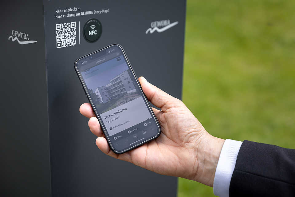 Eine Person hält ein Smartphone in der Hand, das die Webseite eines Bauprojekts mit dem Titel 'Tarzan und Jane' zeigt. Das Smartphone wird nahe an eine Infotafel gehalten, die einen QR-Code und ein NFC-Logo enthält. Auf der Tafel steht 'GEWOBA' und sie bietet eine Anleitung zum Abrufen weiterer Informationen über die GEWOBA Story-Map. Im Hintergrund ist ein grüner Rasen zu sehen.