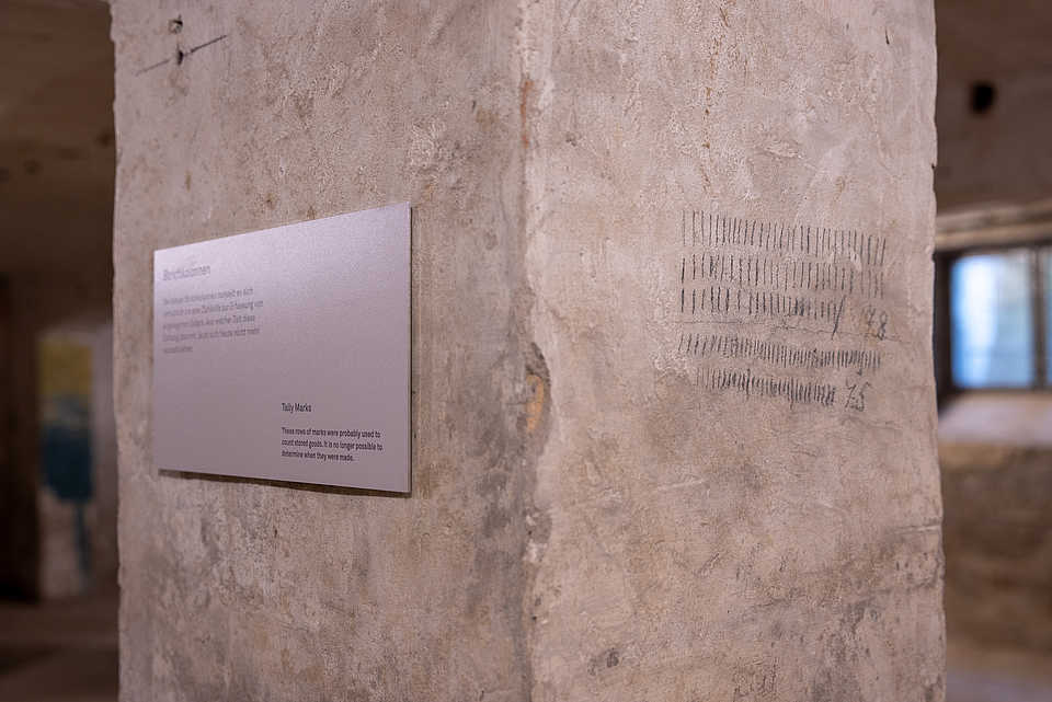 Eine Steinsäule mit eingeritzten Strichlisten. Daneben ist eine Metalltafel mit der Überschrift "Strichlisten" angebracht.