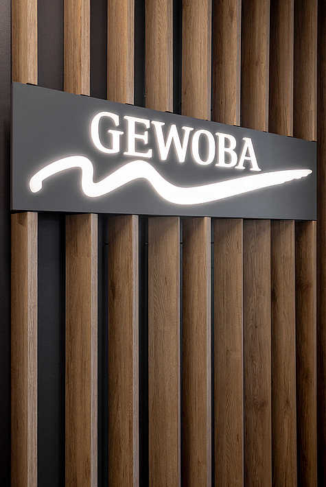 GEWOBA-Logo in Weiß auf dunklem Hintergrund, montiert auf senkrechten Holzlamellen.