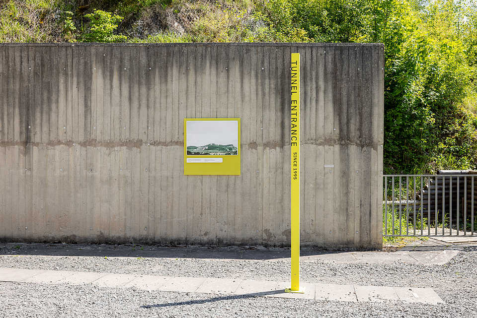 Außenansicht des Tunnelzugangs zur KZ-Gedenkstätte Mittelbau-Dora mit gelber Stele: ‚Tunnel Entrance – Since 1995‘ und Infotafel an der Betonwand.