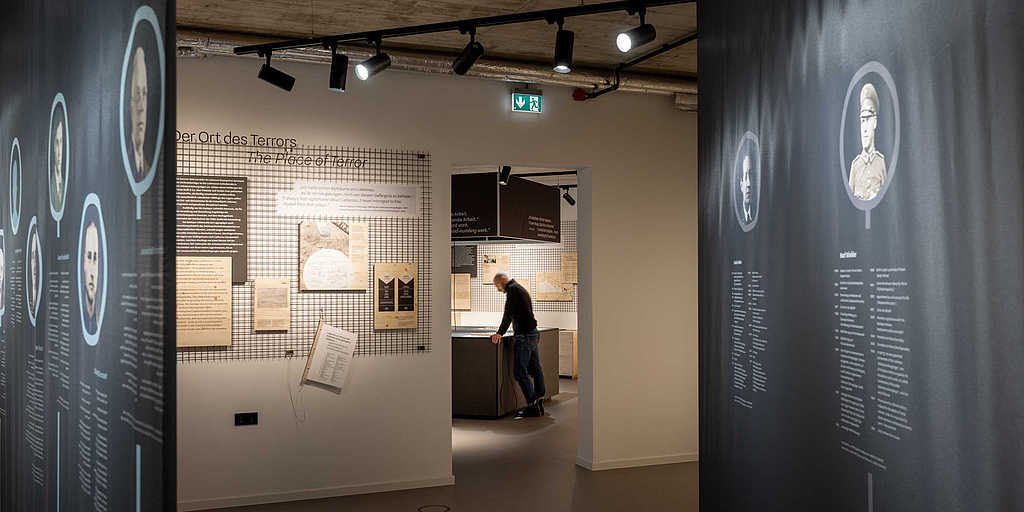 Ausstellungsraum mit Porträts auf Stoffbahnen und Informationstafeln an einer Gitterwand.
