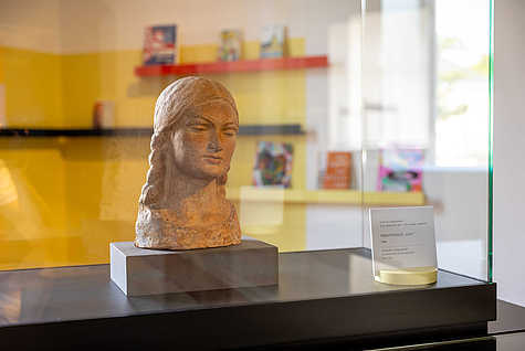 Eine Büste aus Terrakotta, die den Kopf eines jungen Mädchens darstellt, wird in einer gläsernen Vitrine ausgestellt. Die Skulptur hat geflochtenes Haar und einen ruhigen Gesichtsausdruck. Daneben steht eine Beschilderung mit dem Titel 'Mädchenkopf - Leni'. Im Hintergrund sind verschwommen Bücherregale und gelbe Wände zu erkennen.