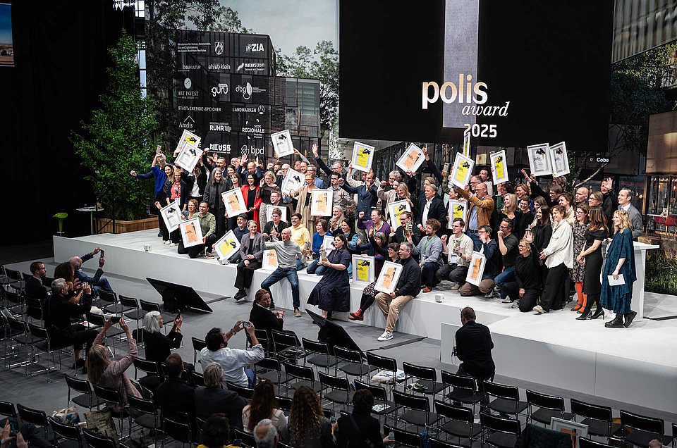Seitliche Aufnahme der preisgekrönten Gruppen auf der Bühne des polis award 2025. Viele Teilnehmer:innen halten Auszeichnungen hoch. Im Vordergrund Publikum, das Fotos macht.