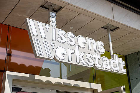Weißes Schild mit dem Logo der Wissenswerkstadt Bielefeld über dem Eingang, mit einer modernen Gestaltung aus Linien und Punkten.
