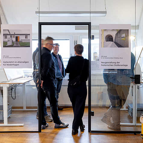 Vier Personen stehen in einem Ausstellungsraum und unterhalten sich, während sie durch eine Glastür sichtbar sind. Links und rechts der Tür sind Poster mit Informationen zu Gedenkorten und Neugestaltungen historischer Anlagen angebracht.