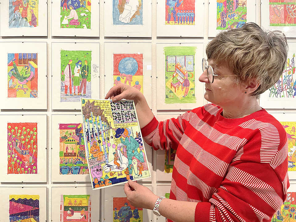 Eine Frau mit kurzem Haar und Brille trägt einen rot-weiß gestreiften Pullover und steht vor einer Wand mit bunten Illustrationen. Sie hält eine Zeichnung in beiden Händen und betrachtet sie aufmerksam. Die Illustrationen im Hintergrund sind ebenfalls farbenfroh und zeigen verschiedene Szenen und Figuren.