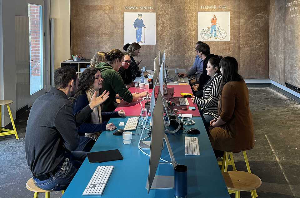 Mehrere Menschen sitzen an einem langen Arbeitstisch und arbeiten an Computern. An der Wand hängen zwei Poster mit den Figuren Ava und Günther. Die Atmosphäre wirkt konzentriert und kollaborativ.