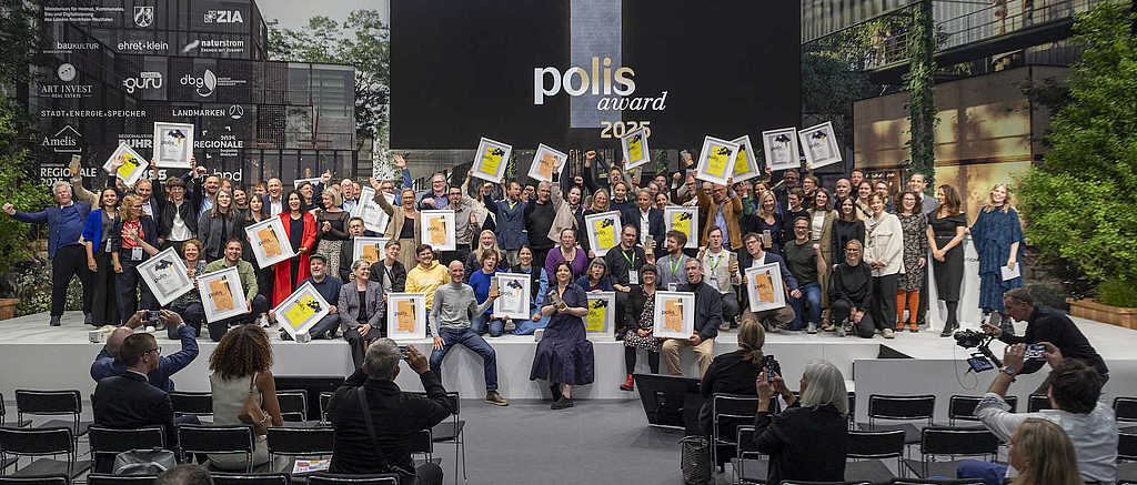Große Gruppenaufnahme aller Gewinner:innen des polis award 2025 auf der Bühne des polis FORUM. Zahlreiche Personen halten gerahmte Urkunden hoch. Im Hintergrund eine große Leinwand mit dem Schriftzug ‚polis award 2025‘.