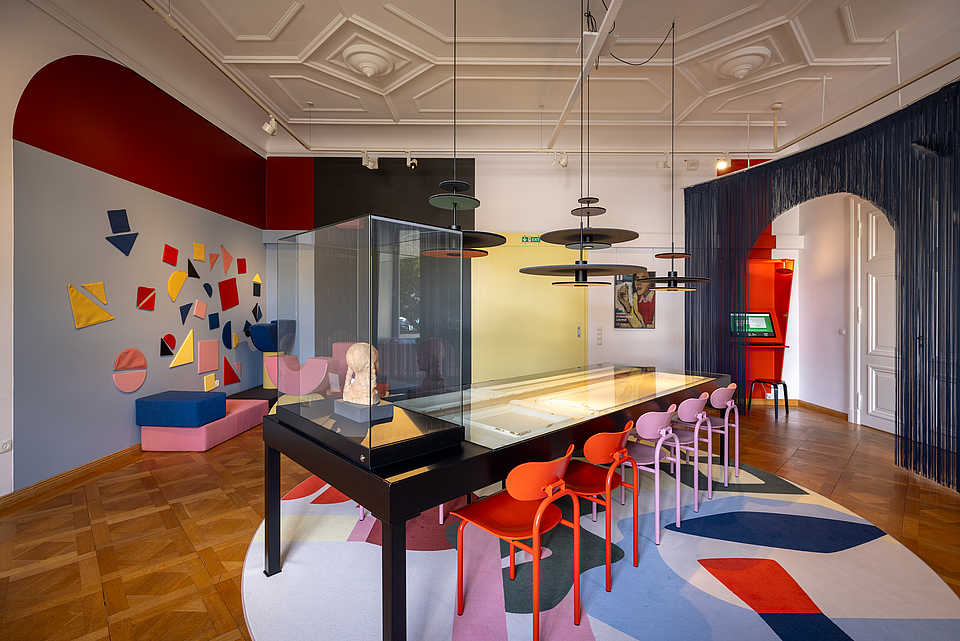 Ein moderner, bunter Raum mit einer Vitrine in der Mitte, die ein Kunstwerk ausstellt. Um die Vitrine herum stehen bunte Stühle in Rot, Pink und Lila an einem großen Tisch. Die Wände sind mit farbigen geometrischen Formen dekoriert, und im Hintergrund sind moderne Hängeleuchten zu sehen. Der Raum hat eine Mischung aus roten und gelben Akzenten sowie eine Decke mit dekorativen Elementen, die den Raum hell und offen wirken lassen.