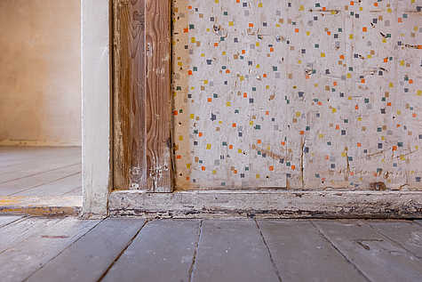 Nahaufnahme einer Wand mit farbigem Tapetenmuster in einem Raum. Der untere Bereich zeigt einen Türrahmen aus Holz sowie einen abgenutzten Holzboden.