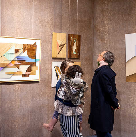 Drei Personen, darunter ein Kind, betrachten geometrische Kunstwerke aus Holz an einer Ausstellungswand. Die Werke sind in warmen Holztönen mit farbigen Akzenten gestaltet.