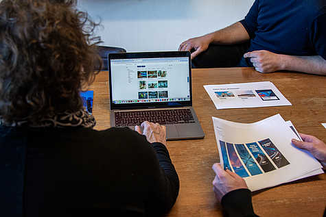 Zwei Personen sitzen an einem Tisch, eine Frau bedient einen Laptop mit geöffneter Bilddatenbank; auf dem Tisch liegen Ausdrucke von Layouts.