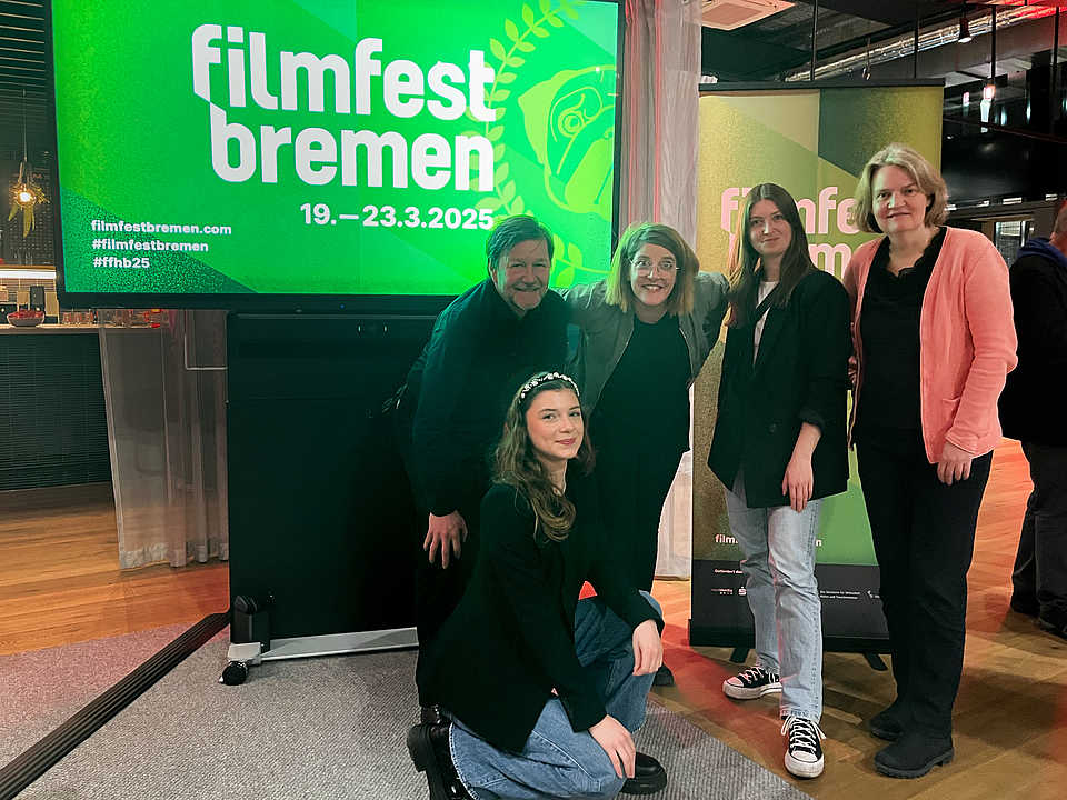 Fünf Teammitglieder der GfG posieren lachend vor einem großen grünen Bildschirm mit dem Logo vom Filmfest Bremen und den Terminen vom 19. bis 23. März 2025.