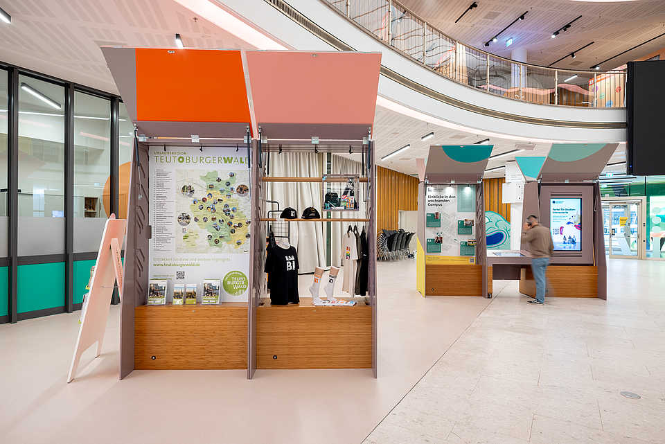 Helle Ausstellungshalle mit farbigen Ständen und einer großen Karte der Region Teutoburger Wald. Die Stände zeigen regionale Highlights und Produkte wie Taschen, T-Shirts und Informationsmaterialien. Im Hintergrund weitere interaktive Ausstellungsbereiche.