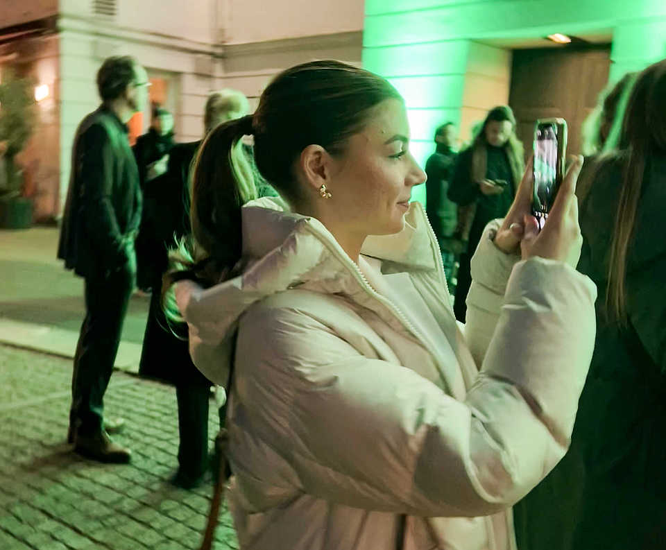 Alea fotografiert mit dem Smartphone vor dem beleuchteten Theater am Goetheplatz bei Nacht. Die Szenerie ist in grünem Licht getaucht, Menschen stehen im Hintergrund.