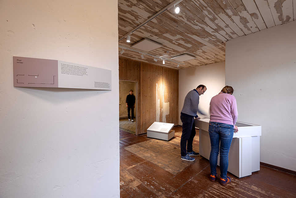 Zwei Personen betrachten eine Ausstellungstafel mit Text und Bildern. Im Hintergrund befindet sich ein weiterer Raum mit einer beleuchteten Wandtafel.