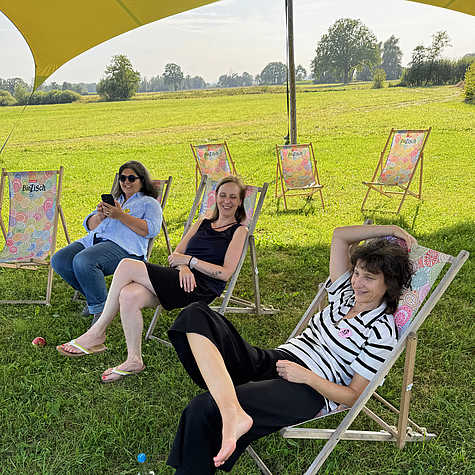 Drei Frauen entspannen auf bunten Liegestühlen unter einem Sonnensegel in einer grünen Wiese. Im Hintergrund sind weitere Liegestühle und eine weite Landschaft zu sehen.