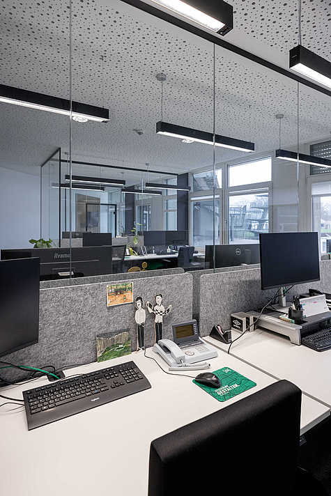 Arbeitsplatz mit weißen Schreibtischen, grauen Trennwänden und mehreren Monitoren. Auf dem Tisch liegen Tastatur, Telefon, Büromaterialien und kleine Dekorationen. Im Hintergrund große Fensterfront.