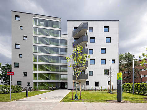 Ein modernes Wohngebäude mit zwei unterschiedlichen Fassaden: links eine graue Fassade mit großen, grünen Fenstern und rechts eine weiße Fassade mit Balkonen. Der Eingangsbereich ist durch einen gepflasterten Weg erreichbar, der von grünen Rasenflächen und jungen Bäumen flankiert wird. Ein Schild mit der Aufschrift 'Feuerwehrzufahrt' und eine Informationssäule zur 'Gartenstadt Vahr' sind ebenfalls zu sehen. Der Himmel ist bewölkt.