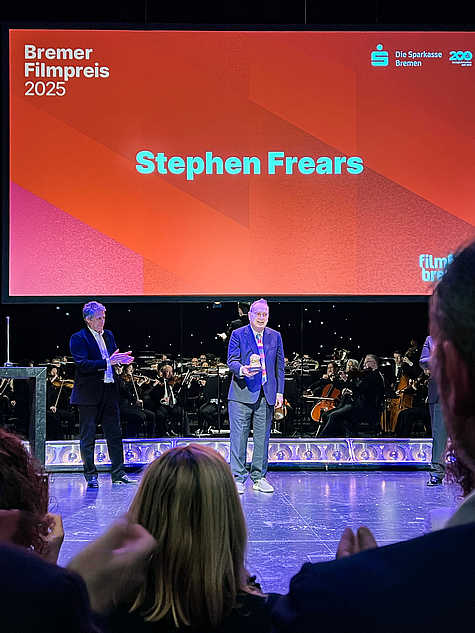 Stephen Frears steht auf der Bühne mit dem Bremer Filmpreis in der Hand. Im Hintergrund spielt ein Orchester. Auf der großen Leinwand über ihm steht „Stephen Frears – Bremer Filmpreis 2025“.