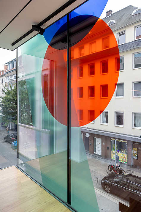 Ein Blick durch bunte Glasflächen mit roten und blauen Kreisen, die auf die Straße und umliegende Gebäude projiziert werden.