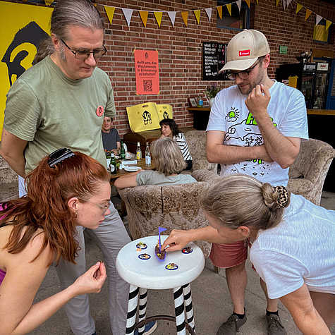 Gruppe von Personen, die sich um einen kleinen Tisch versammelt haben und ein Spiel mit kleinen Figuren spielen. Im Hintergrund sitzen weitere Menschen auf Sofas vor einer Ziegelfassade mit gelber Dekoration und Bannern.