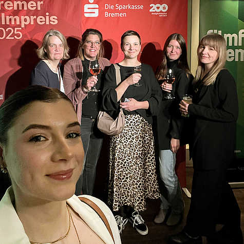 Alea macht ein Selfie von sich und ihren Kolleginnen vor einem roten Hintergrund mit dem Logo der Sparkasse Bremen und dem Schriftzug "Bremer Filmpreis 2025".