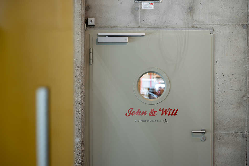 Ein Durchgang mit zwei alten, gelben Metalltüren führt zu einer modernen Tür mit der Aufschrift "John & Will" in roter Schrift. Die Wände sind aus Sichtbeton, die gelben Türen zeigen Gebrauchsspuren.
