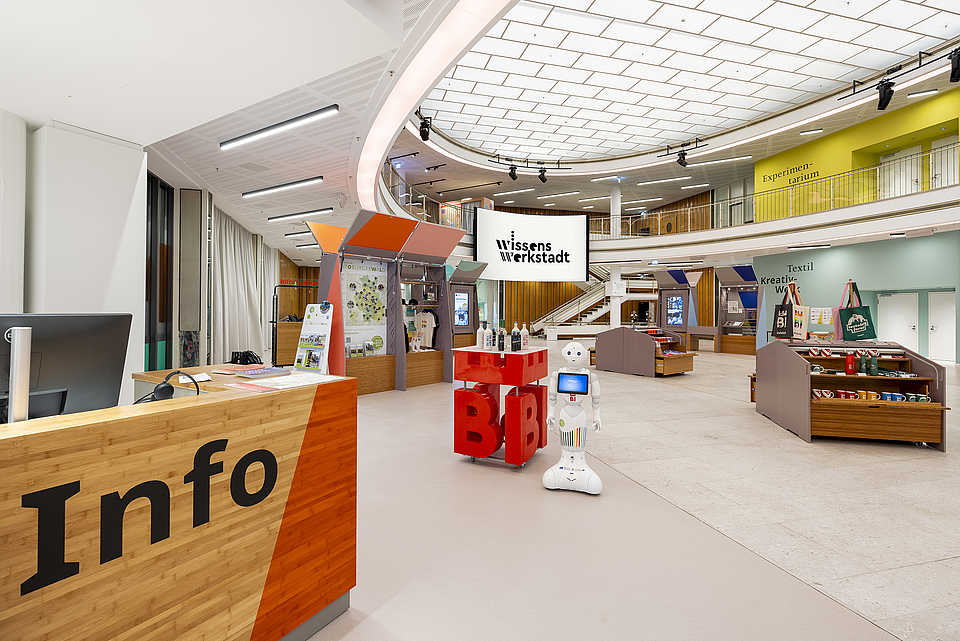 Ein heller Empfangsbereich mit einem großen Informationsschalter aus Holz. Im Hintergrund sind bunte Ausstellungsmodule, ein humanoider Roboter und ein großer Bildschirm mit dem Logo der Wissenswerkstatt zu sehen.