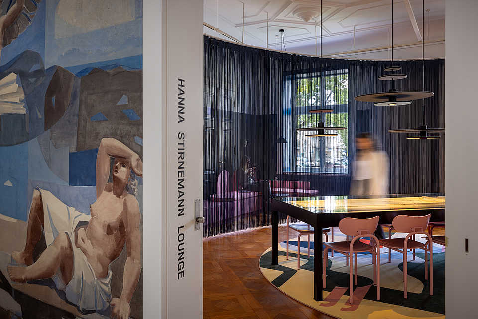 Eingang zur 'Hanna Stirnemann Lounge', dekoriert mit einem Wandgemälde eines sitzenden, halbnackten Mannes. Der Raum ist durch eine Vorhang-Abtrennung geteilt, mit rosa Stühlen um einen langen Tisch und modernen Hängeleuchten. Im Hintergrund sitzt eine Person auf einer rosafarbenen Couch und arbeitet. Eine unscharf abgebildete Person geht an einem beleuchteten Tisch vorbei.