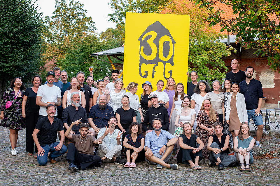 Das Bild zeigt eine große Gruppe von Menschen, die für ein Gruppenfoto vor einem gelben Hintergrund mit der Aufschrift "30 GfG" posieren. Die Personen wirken fröhlich und entspannt. Im Hintergrund sind Bäume und ein Gebäude sichtbar.