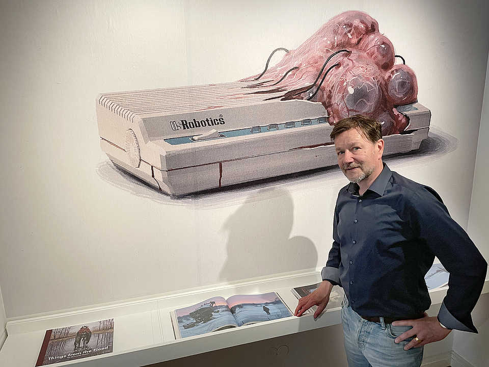Ein Mann mit dunklem Hemd und Jeans steht vor einer Wandinstallation, die eine skulpturale Darstellung eines Geräts mit der Aufschrift 'Robotics' zeigt, das teilweise von organisch aussehenden Formen bedeckt ist. Er blättert in einem Buch auf einem weißen Regal, das sich unter der Installation befindet. Die Szene ist in einer Galerie oder einem Ausstellungsraum aufgenommen.