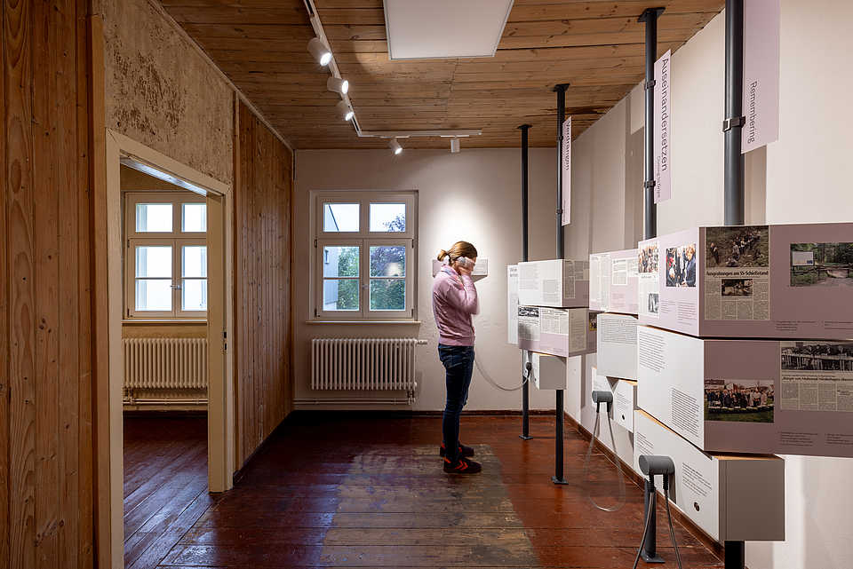 Eine Person steht in einem Raum mit hölzernem Boden und vertäfelter Decke, umgeben von Tafeln mit Ausstellungsinformationen.