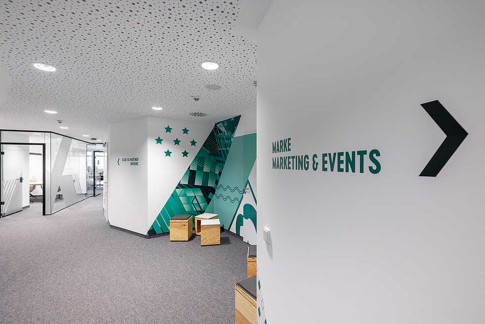 Blick auf einen Büroflur. An der weißen Wand rechts zeigt ein Pfeil den Weg zur Abteilung ‚Marke, Marketing & Events‘. Links führen Glaswände zu weiteren Büroräumen.“