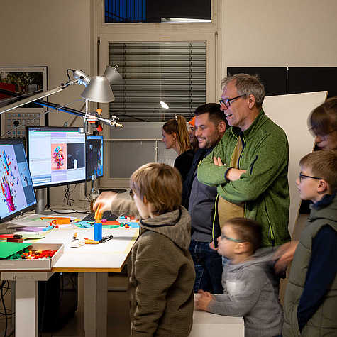 Das Bild zeigt eine Gruppe von Erwachsenen und Kindern, die sich an einem Computerarbeitsplatz versammelt haben. Auf den Monitoren sind kreative Inhalte zu sehen, und auf dem Tisch davor liegen bunte Bausteine und Bastelmaterial.