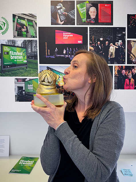 Frau in einem Ausstellungsraum küsst eine goldene Skulptur in Form eines Hundekopfes. Im Hintergrund sind Plakate und Fotos zum Filmfest Bremen 2024 an der Wand zu sehen.
