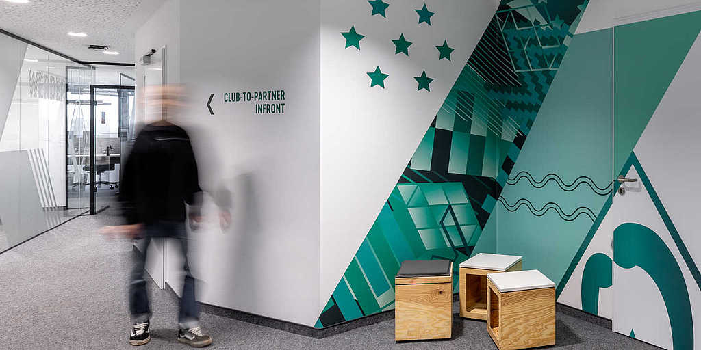 Flur im Bürogebäude mit Wandgrafiken in Grün- und Türkistönen, Sternen und dem Schriftzug ‚MOIN.‘. Links führt ein Glaskorridor zu weiteren Räumen, ein Mensch geht durch das Bild.