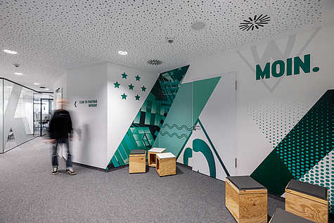 Flur im Bürogebäude mit Wandgrafiken in Grün- und Türkistönen, Sternen und dem Schriftzug ‚MOIN.‘. Links führt ein Glaskorridor zu weiteren Räumen, ein Mensch geht durch das Bild.