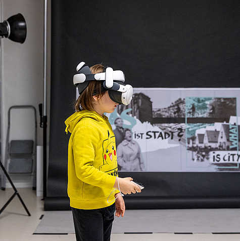 Ein Kind in einem gelben Pikachu-Pullover trägt eine Virtual-Reality-Brille und hält einen Controller, während es vor einem schwarzen Hintergrund mit Collagen steht.