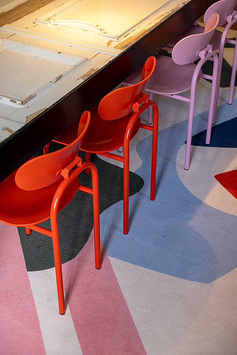 Nahaufnahme von bunten Stühlen in Rot und Rosa, die um einen großen Tisch mit einer Glasplatte angeordnet sind. Der Boden ist mit einem Teppich in geometrischen Mustern in Blau, Rosa und Grau bedeckt, was dem Raum eine lebhafte Atmosphäre verleiht.