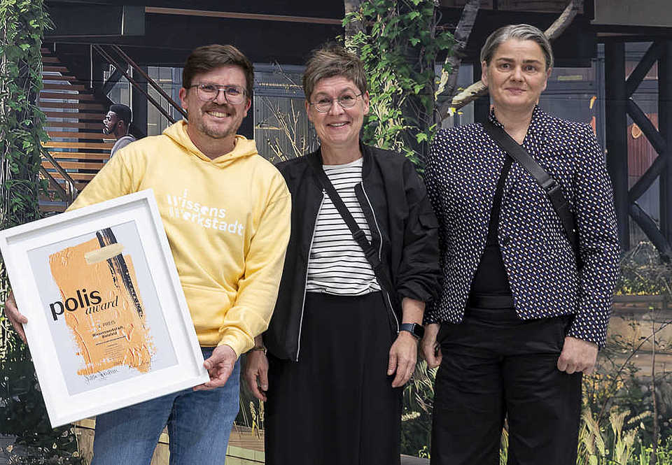 Drei Personen stehen vor einem Architektur-Poster. Links hält eine Person im gelben Hoodie eine gerahmte polis-award-Urkunde für die Wissenswerkstadt Bielefeld. Alle drei lächeln in die Kamera.