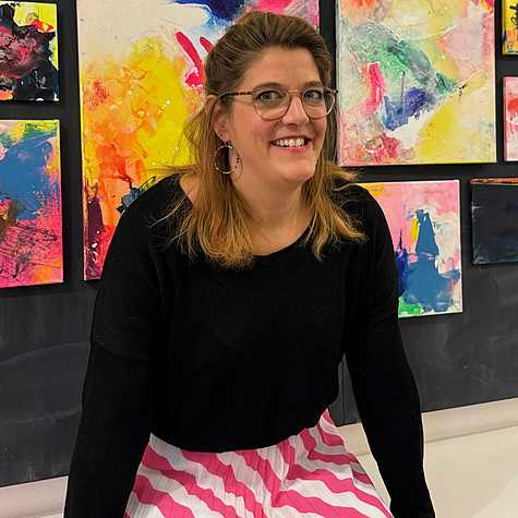 Das Bild zeigt eine lächelnde Frau vor einer Wand mit bunten, abstrakten Kunstwerken. Sie trägt ein schwarzes Oberteil und einen Rock mit pink-weißem Streifenmuster.