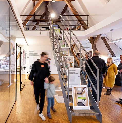 Moderner Ausstellungsraum mit Holzdetails und einer Metalltreppe, die zu einer Galerie führt. Besucher:innen betrachten Infotafeln und Exponate, während eine Frau mit einem Kind durch den Raum geht.