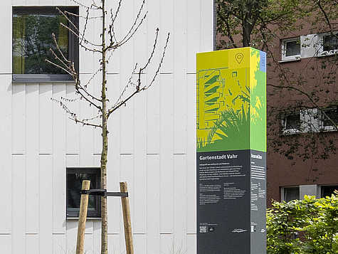 Eine Informationssäule zur 'Gartenstadt Vahr' steht vor einer weißen Gebäudefassade. Die Säule ist in den Farben Grün und Gelb gehalten und enthält Texte sowie eine Karte des Wohngebiets. Neben der Säule steht ein junges, frisch gepflanztes Bäumchen, das von Holzpfählen gestützt wird. Im Hintergrund sind weitere Gebäude und grüne Büsche zu sehen.