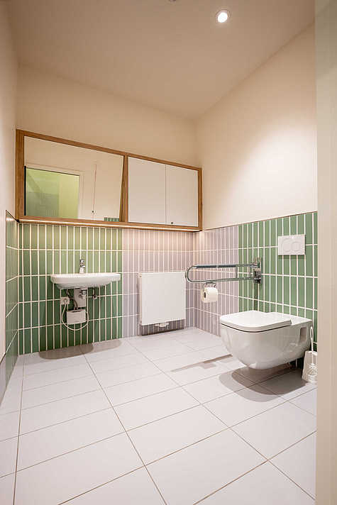 Modernes, barrierefreies Kunden-WC mit grünen Wandfliesen und Haltegriffen.