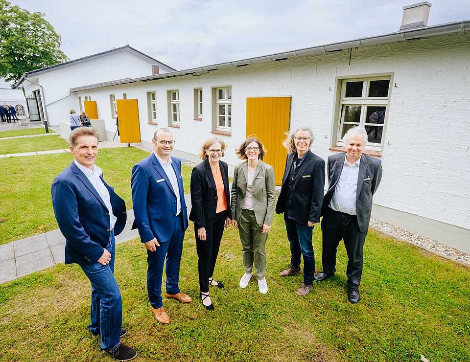 Eine Gruppe von sechs Personen steht vor einem weiß gestrichenen Gebäude mit gelben Türen und Fenstern.
