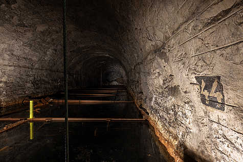 Blick in den dunklen unterirdischen Stollen der KZ-Gedenkstätte Mittelbau-Dora mit Wasser auf dem Boden und rostigen Metallträgern. An der Wand ist die Zahl 44 aufgemalt.