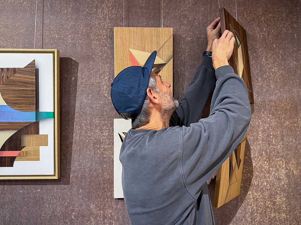 Ein Mann mit Baseballcap hängt ein geometrisches Kunstwerk aus Holz an eine Wand. Im Hintergrund sind weitere Kunstwerke mit ähnlichem Stil sichtbar.
